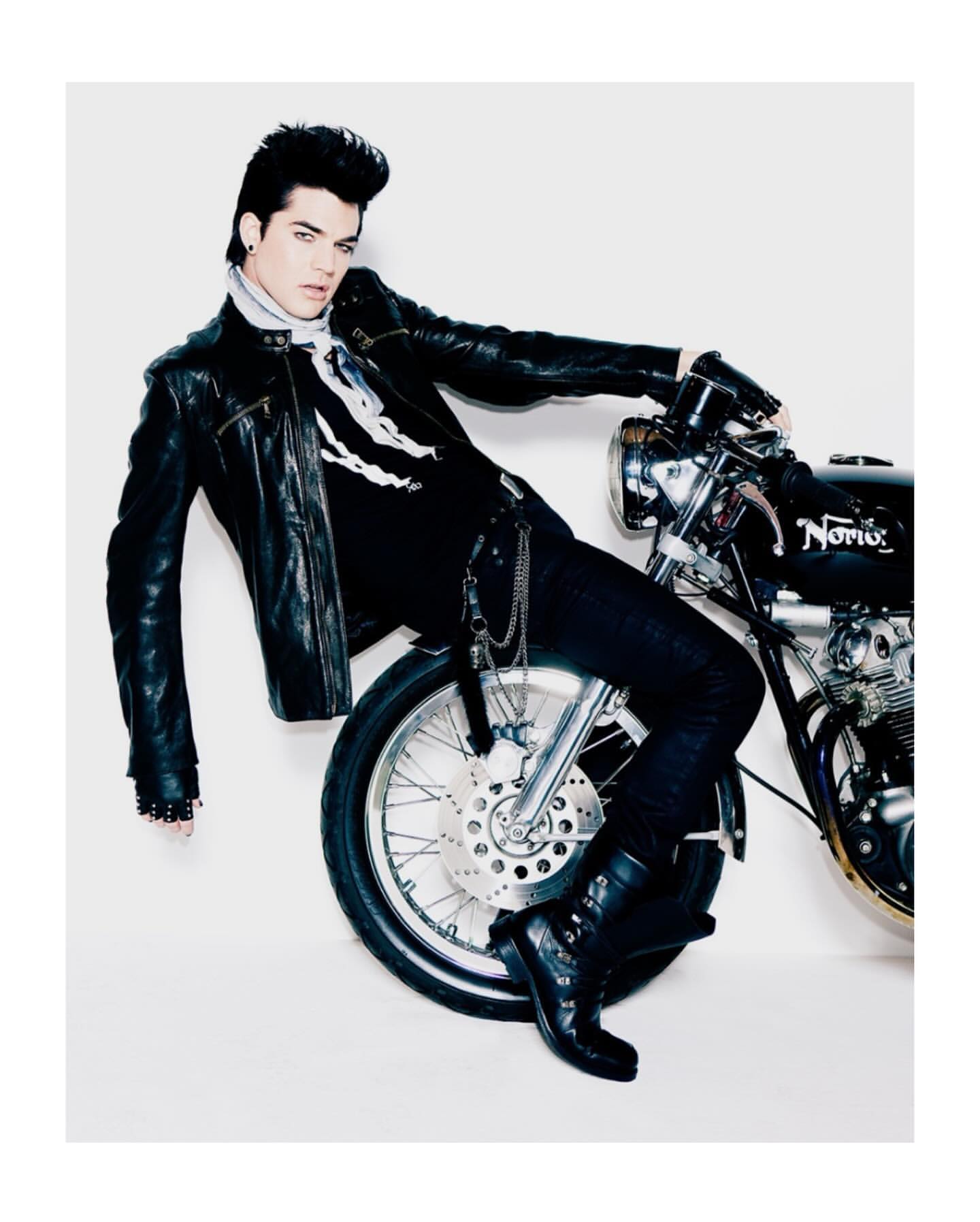 Adam Lambert, singer, poses with a classic black motorcycle in a studio shot, wearing black leather jacket, dark denim, and motorcycle boots.