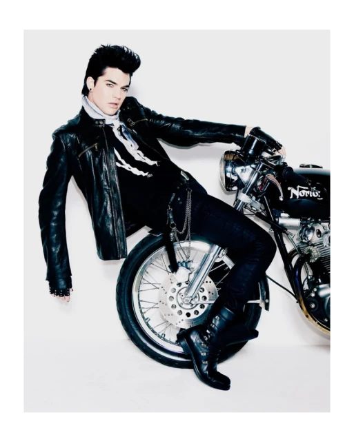 Adam Lambert, singer, poses with a classic black motorcycle in a studio shot, wearing black leather jacket, dark denim, and motorcycle boots.