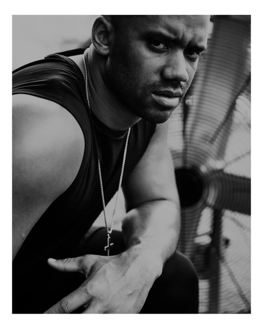Monochrome portrait of a muscular man with a determined expression, wearing a tank top and silver cross necklace, with a fan in the background.
