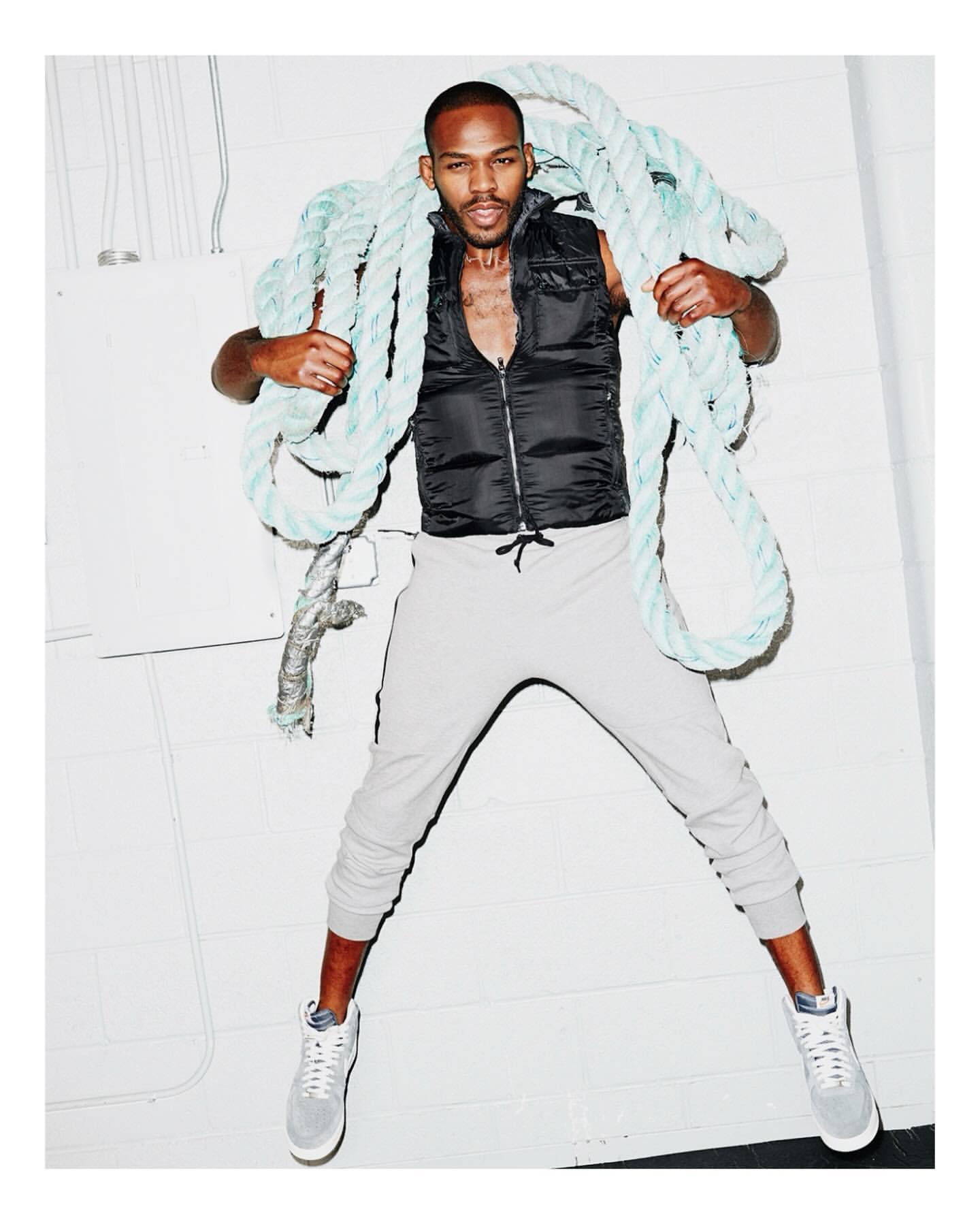 Adult male fashion model poses in gray sweatpants, a black vest, and sneakers while holding a blue rope in a photo studio.