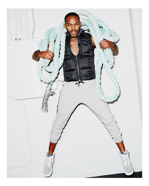 Adult male fashion model poses in gray sweatpants, a black vest, and sneakers while holding a blue rope in a photo studio.