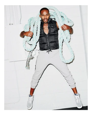 Adult male fashion model poses in gray sweatpants, a black vest, and sneakers while holding a blue rope in a photo studio.