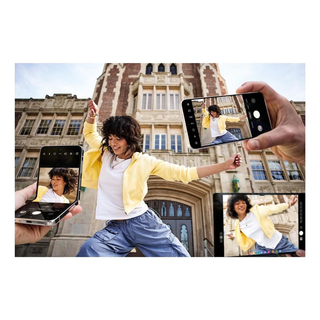 Young woman in blue pants and yellow jacket dances with arms outstretched, framed by hands holding smartphones capturing the moment.