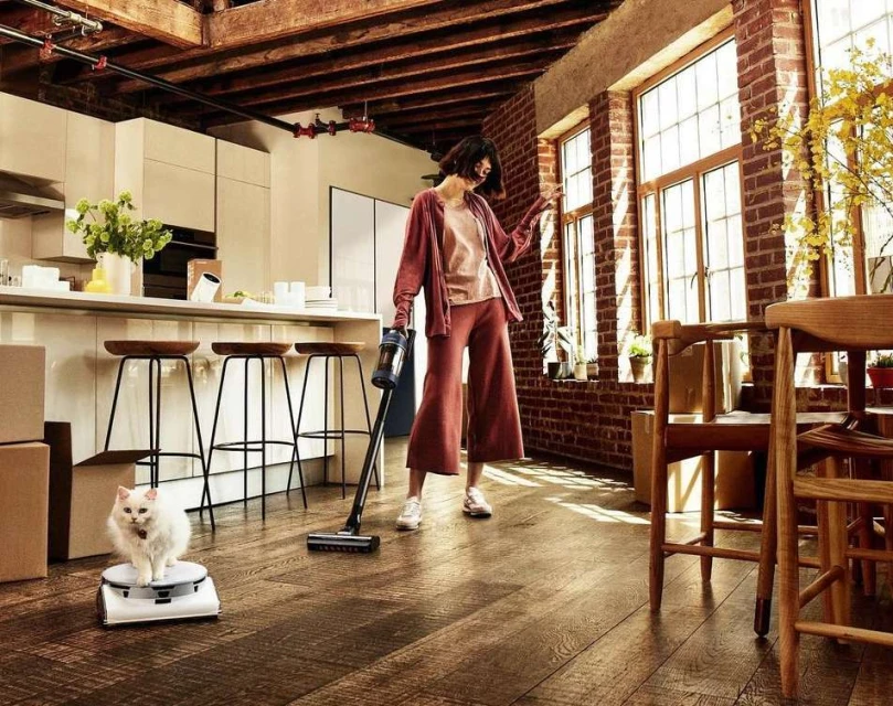 Woman vacuums hardwood floor in a modern apartment; a white cat sits on a scale. Brick walls and natural light fill the space.