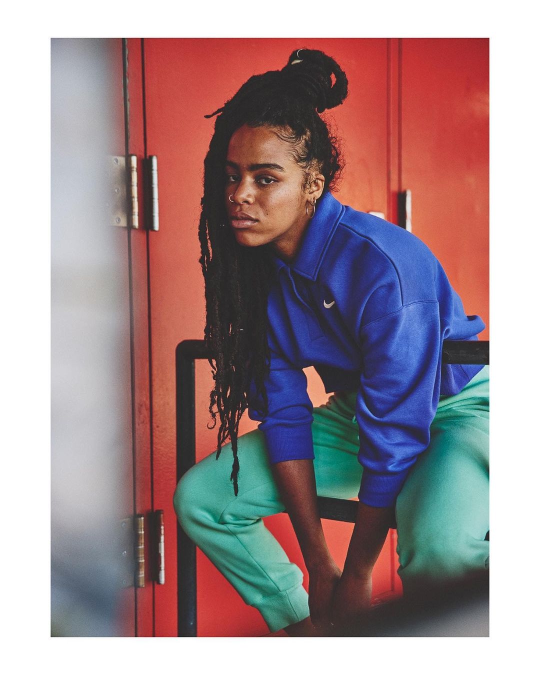 Adult female with long dreadlocks, wearing a blue collared sweater and mint green sweatpants, crouches on black metal bars indoors.