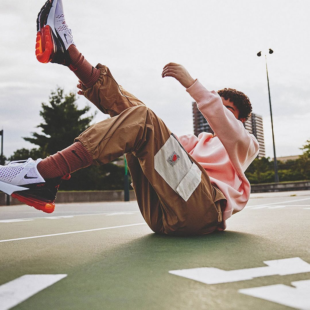 Person in pink sweatshirt and tan pants sitting on a court with white sneakers and orange accents, posing for a fashion shot.
