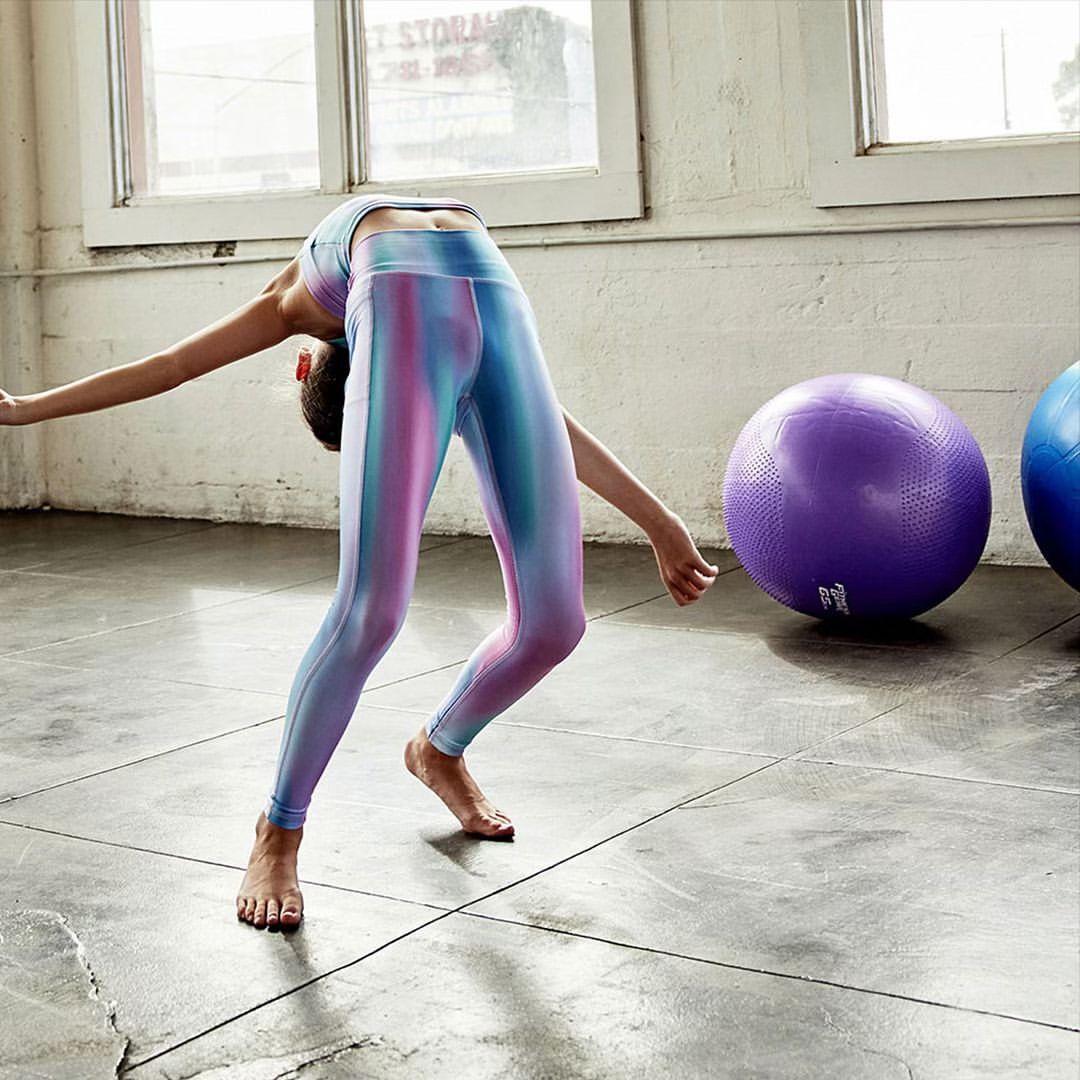 Athletic woman in rainbow leggings does a backbend pose in a brightly lit fitness studio, showcasing a blend of style and fitness.