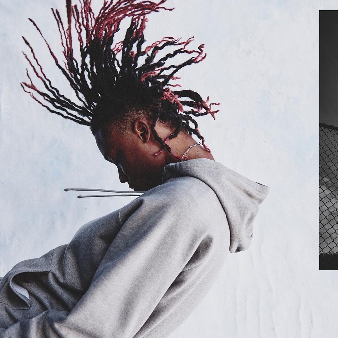 Portrait of a young adult with dreadlocks wearing a gray hoodie, against a white background and partial fence, head tilted down in studio.