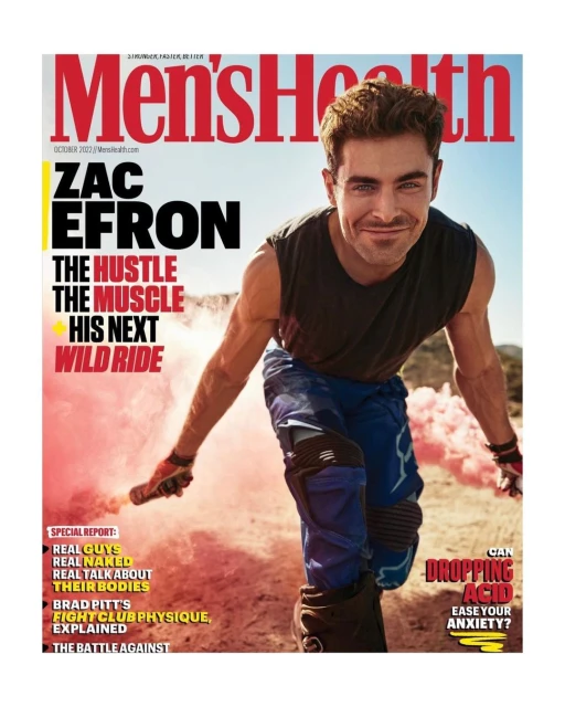 Men's Health magazine cover featuring Zac Efron. Efron poses with pink smoke, wearing a sleeveless shirt and dirt biking pants. Outdoor setting.