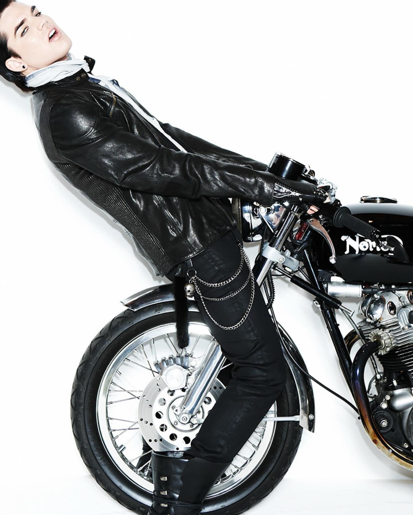 A stylish male model poses with a vintage motorcycle in a studio, wearing a black leather jacket, black jeans, and black boots on a white background.