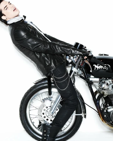 A stylish male model poses with a vintage motorcycle in a studio, wearing a black leather jacket, black jeans, and black boots on a white background.