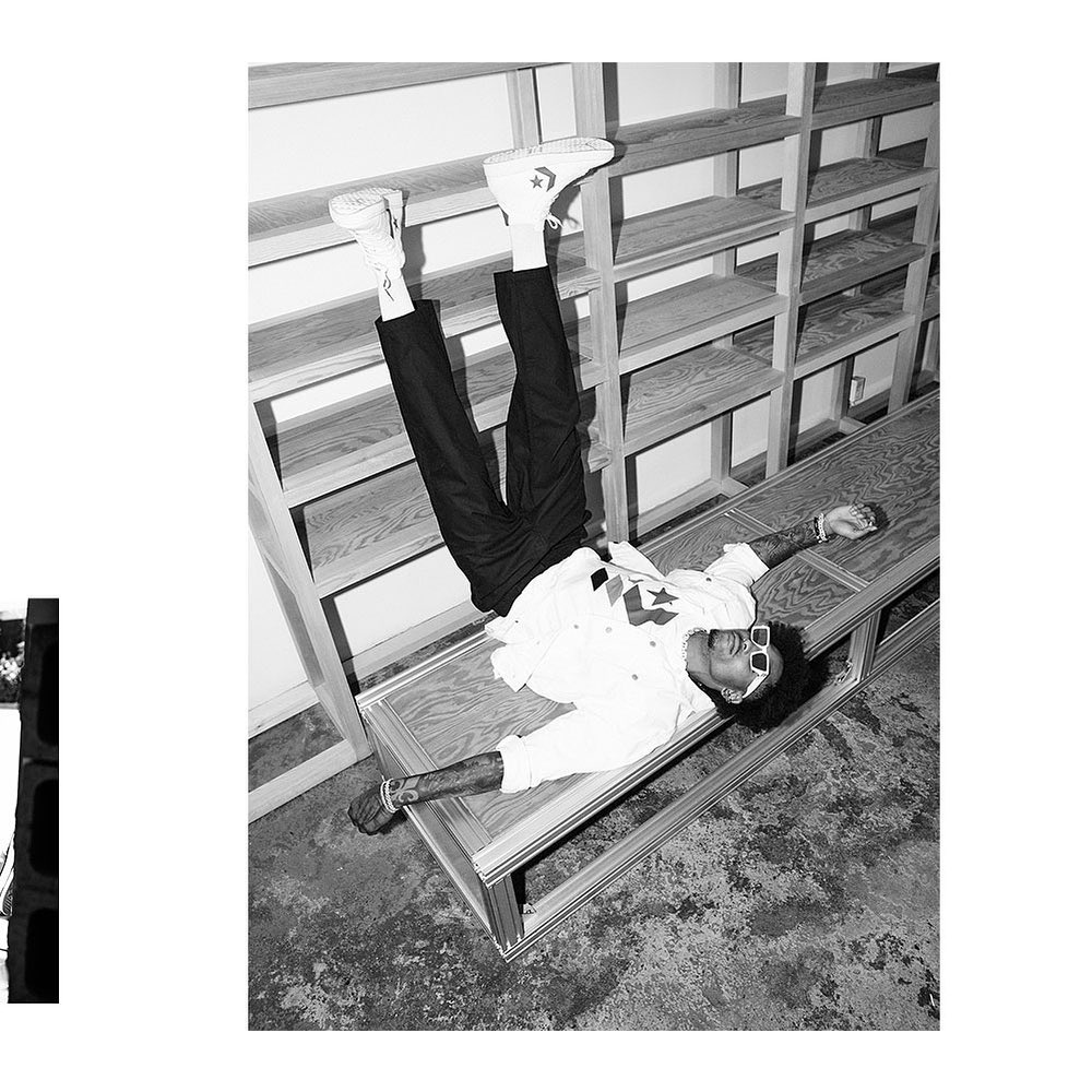 A Black man in a white shirt and black pants lies upside down on a ledge wearing white sneakers and sunglasses in a high-contrast black and white photograph.