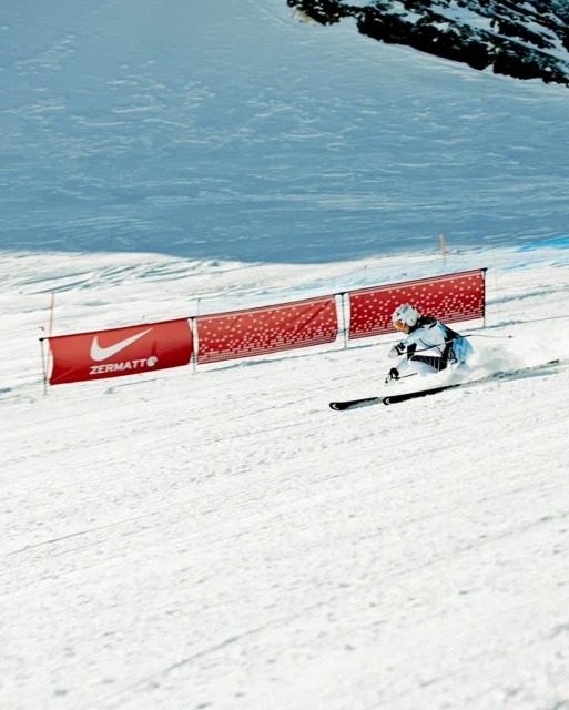A person in ski gear races downhill on a sunny, snow-covered slope with a Nike banner; a competition in the Zermatt region.