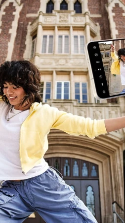 Smiling woman in yellow jacket poses in front of a brick building, reflected in a smartphone camera screen.