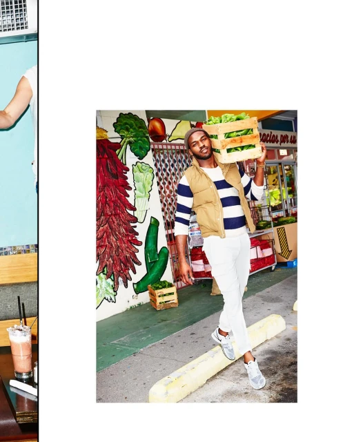 Man in streetwear carries a wooden crate of lettuce in an urban setting with a mural, wearing a vest, striped shirt, and sneakers.