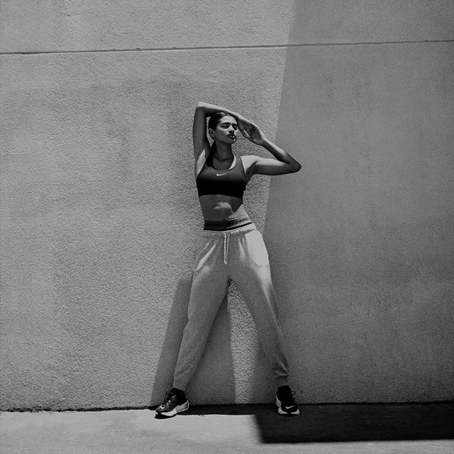 Athletic woman in workout gear poses against a textured wall in black and white. She wears a sports bra and sweatpants, hands raised.