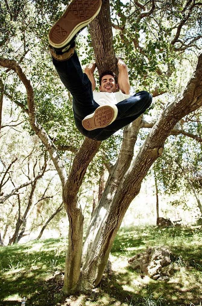 Adult male hangs upside down from a tree branch outdoors wearing casual clothing and sneakers with grass and foliage in the background.