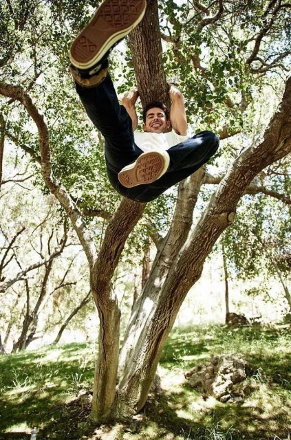 Adult male hangs upside down from a tree branch outdoors wearing casual clothing and sneakers with grass and foliage in the background.