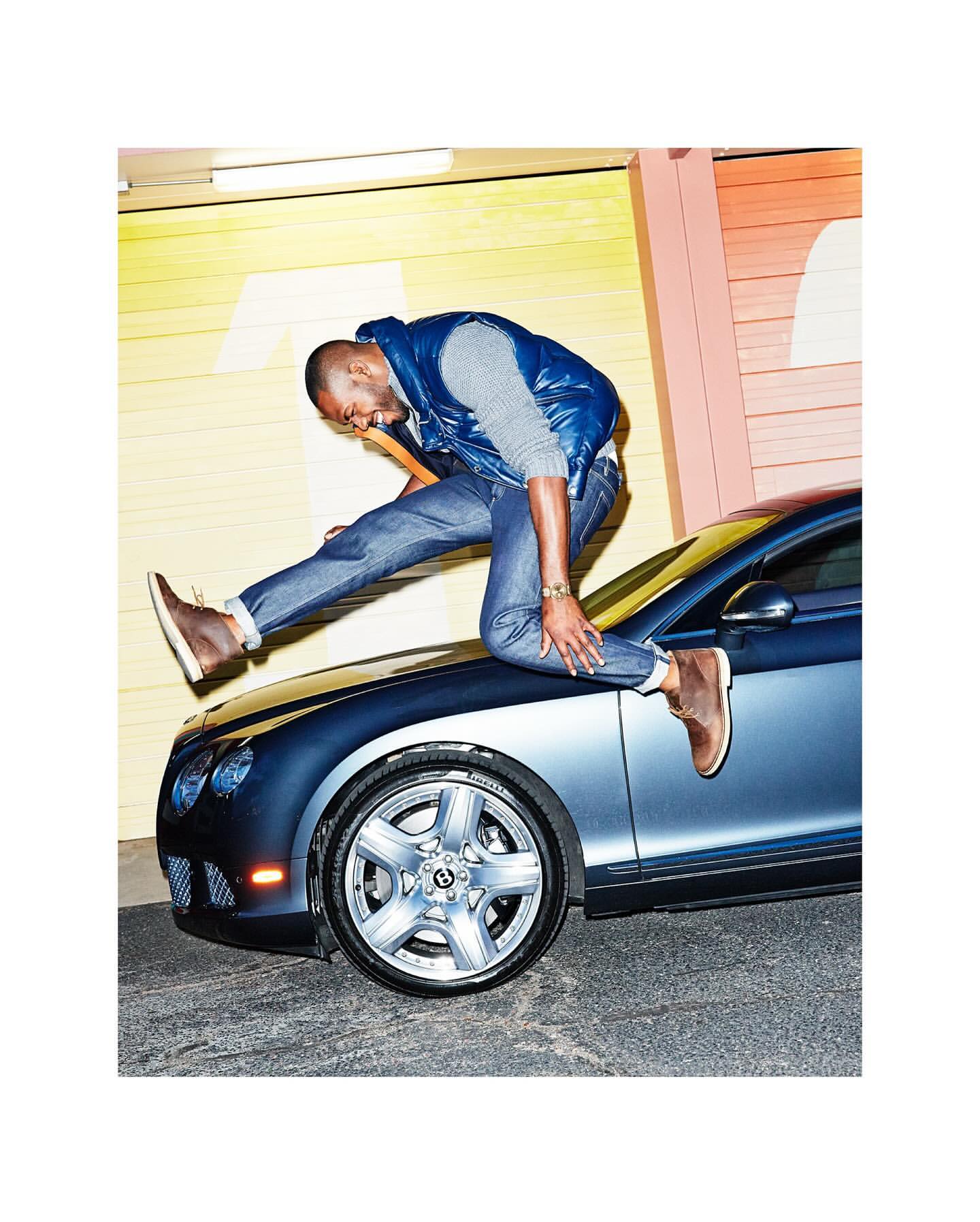 An adult Black man jumping on the hood of a dark blue car in front of a yellow and orange wall, wearing jeans, a vest, and tan shoes.