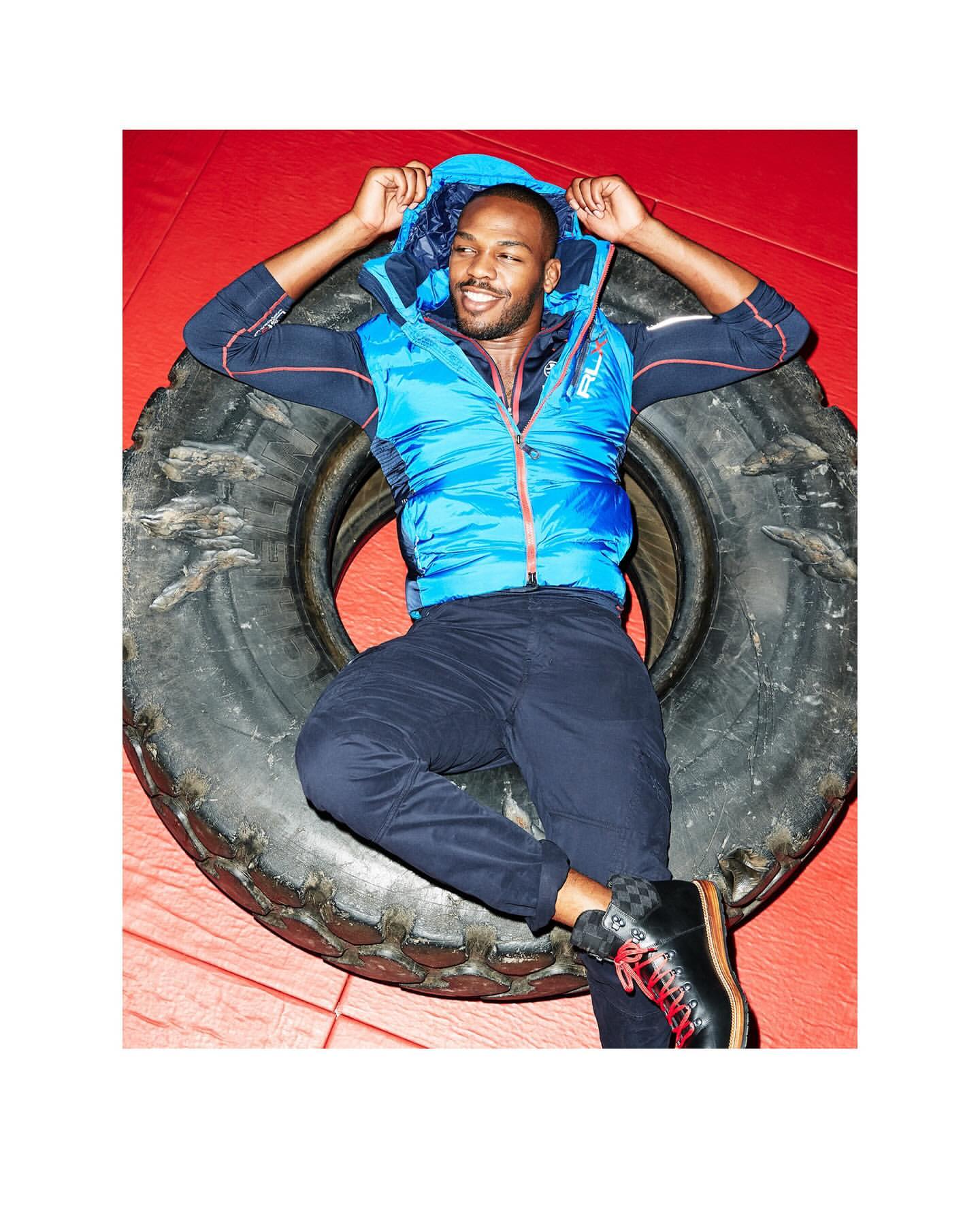 Adult male, wearing a blue vest and dark pants, lying on a black tire in a red indoor setting, smiling and looking at camera.