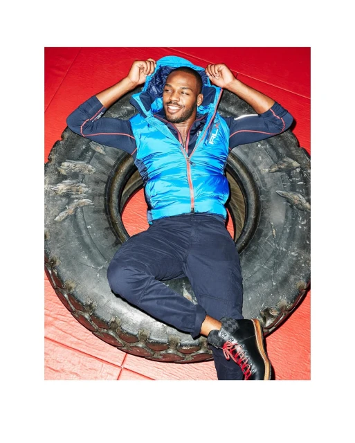 Adult male, wearing a blue vest and dark pants, lying on a black tire in a red indoor setting, smiling and looking at camera.