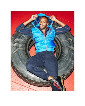 Adult male, wearing a blue vest and dark pants, lying on a black tire in a red indoor setting, smiling and looking at camera.