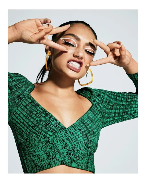 Close-up shot of a young adult woman with bold white eyeliner, gold hoop earrings, and a green patterned crop top making a peace sign.