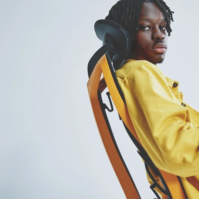 Man with dreadlocks sits in an orange chair wearing a yellow shirt against a white background looking directly at the camera.