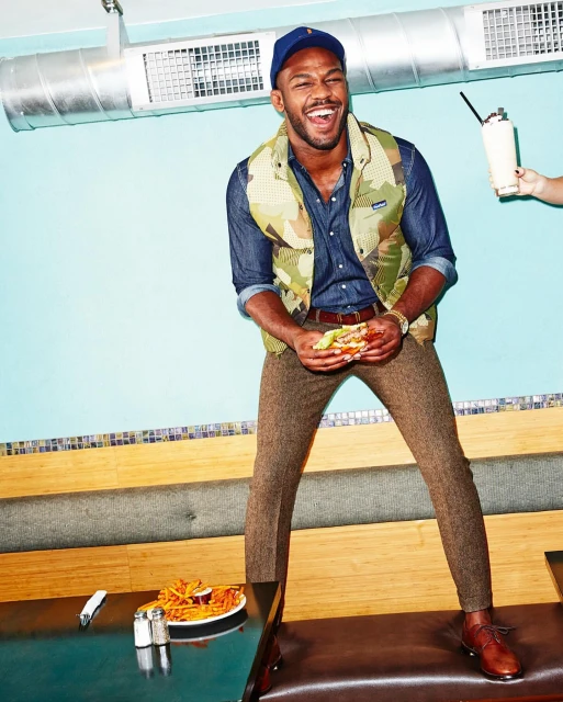 Smiling man in vest and cap stands on bench with burger in hands in diner; fries, drink on table; arm reaches in with milkshake.