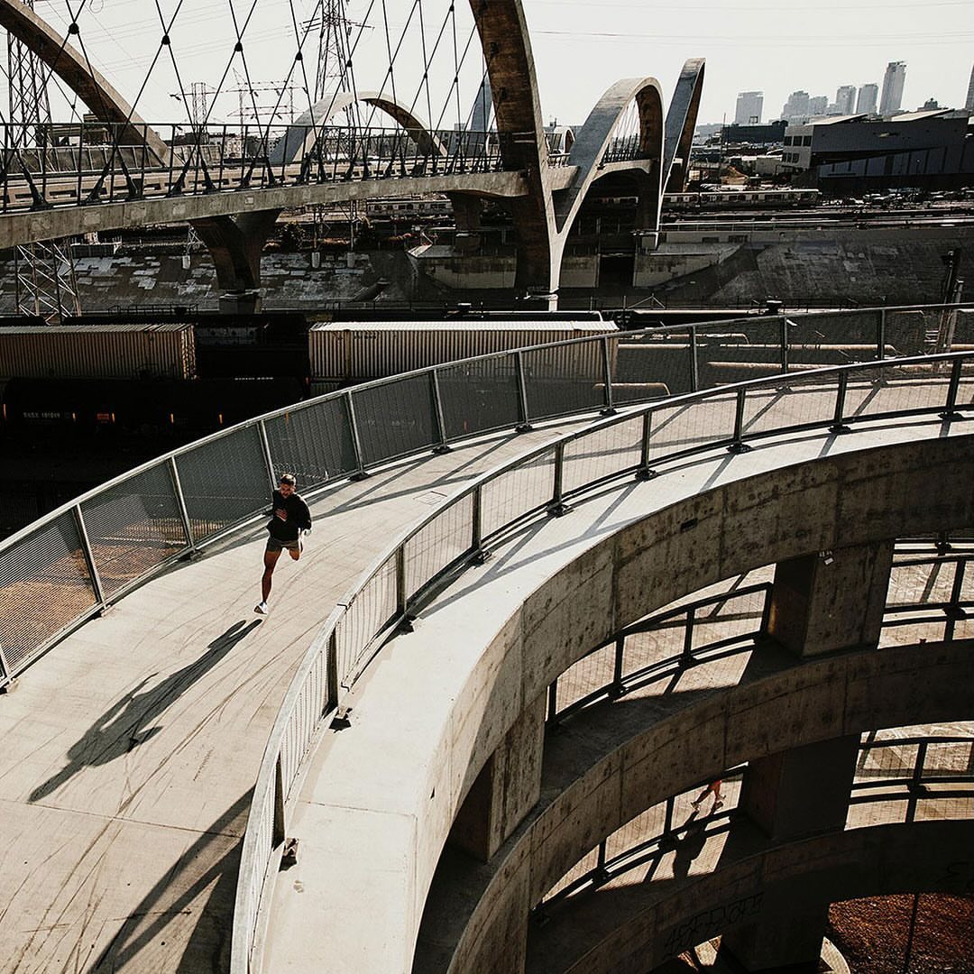An adult runs on a concrete bridge with rounded architectural elements, while a traditional bridge and cityscape rise in the background.