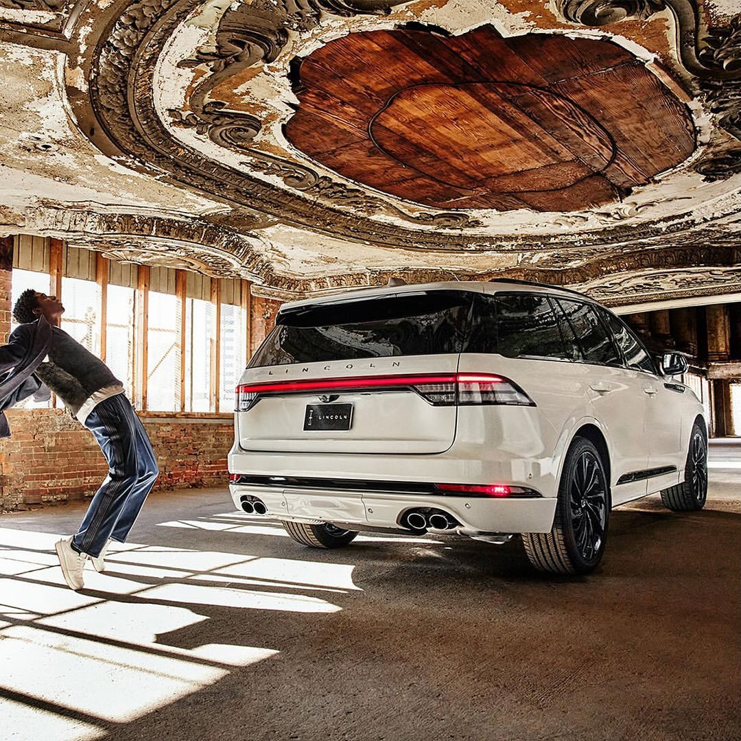 A white Lincoln Aviator SUV is parked indoors with a man leaning against it in a building with peeling paint and decorative ceiling.
