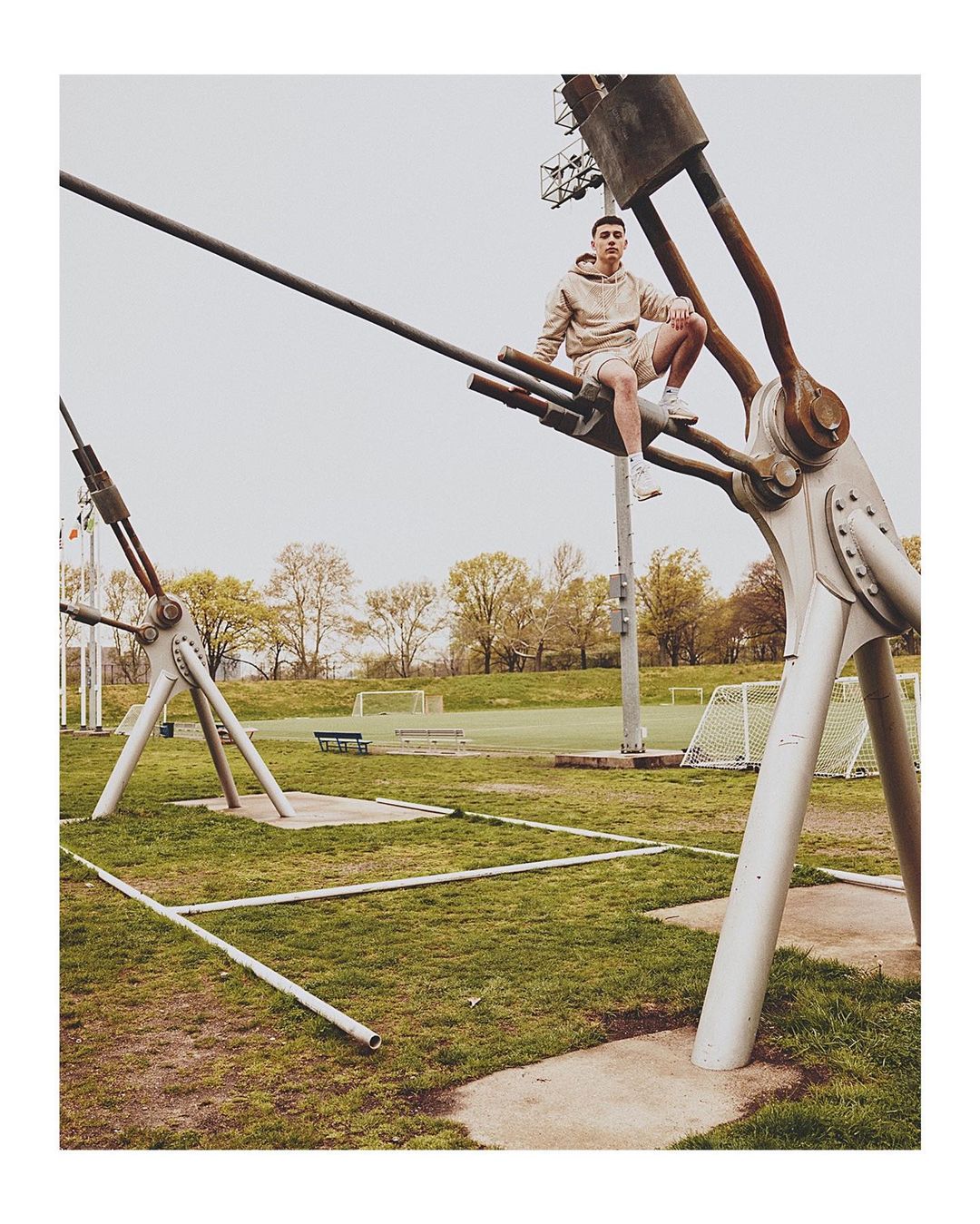 Young man in athletic wear is sitting on metal structure in a green urban park setting with blue sky and trees, shot at eye-level.