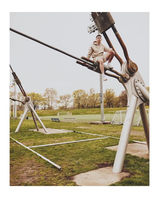 Young man in athletic wear is sitting on metal structure in a green urban park setting with blue sky and trees, shot at eye-level.