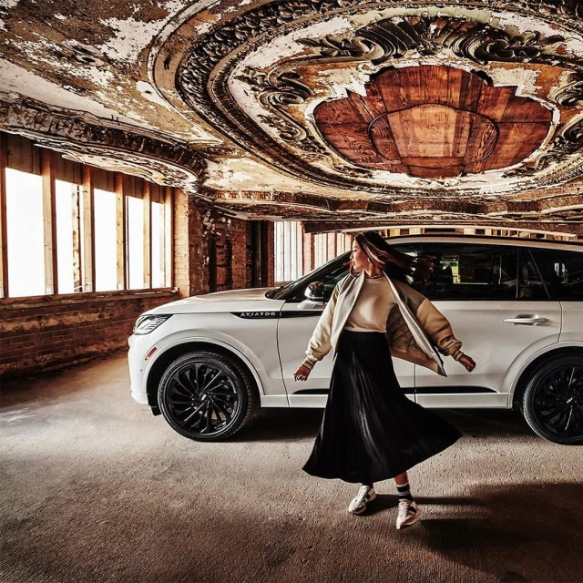 Woman in flowing skirt poses next to a white SUV in a weathered building with ornate ceiling details and tall windows for an automotive ad.