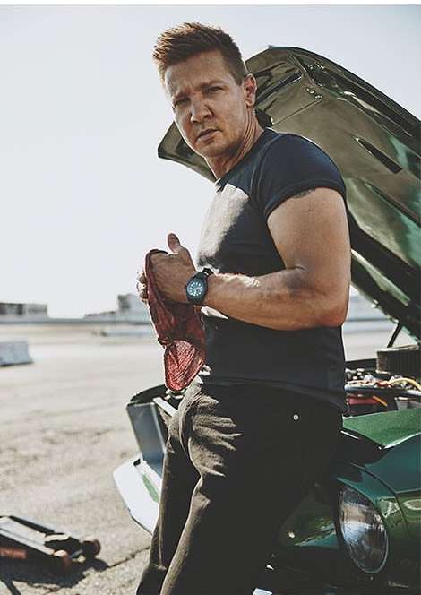 Adult male mechanic leans on an open, dark-green car hood with a wrench, wearing dark work clothes, outdoors on pavement in sunlight.