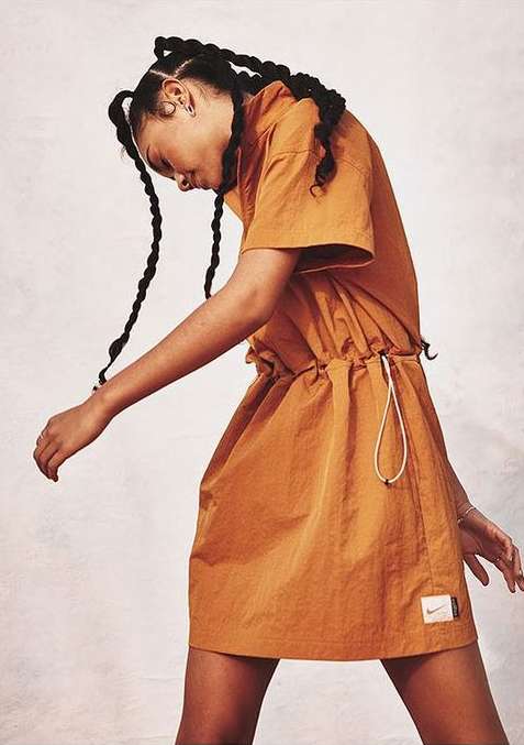 A young woman poses in a caramel-colored Nike dress with braided hair against a soft white background in a studio setting.