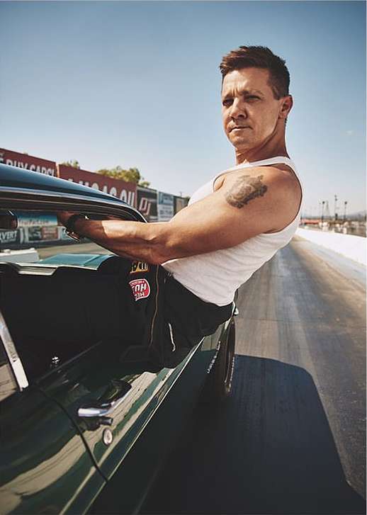 Man leaning out of vintage car on race track, wearing a white tank top. Blue sky and grandstands in background, vintage car focus.