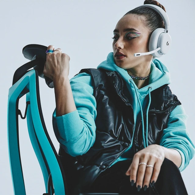 A woman with light skin poses wearing a black vest, turquoise hoodie, gaming headset, and rings while sitting in a turquoise chair.