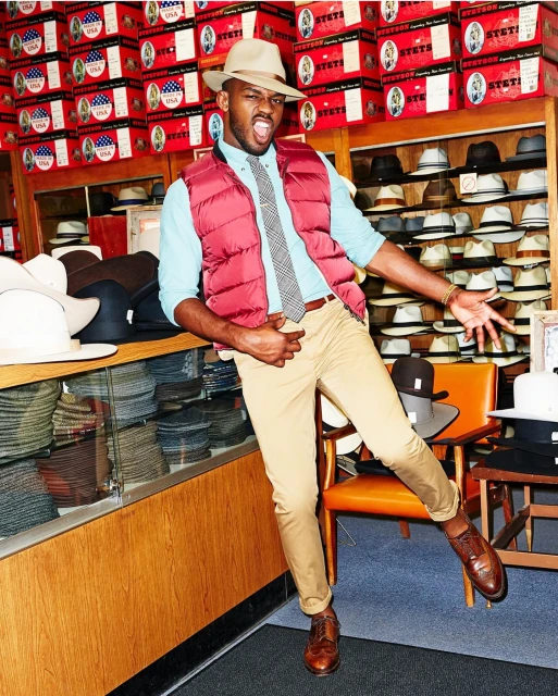 Stylish adult male in hat shop striking a pose, wearing a salmon vest and tan pants, surrounded by hat boxes and display shelves of hats.