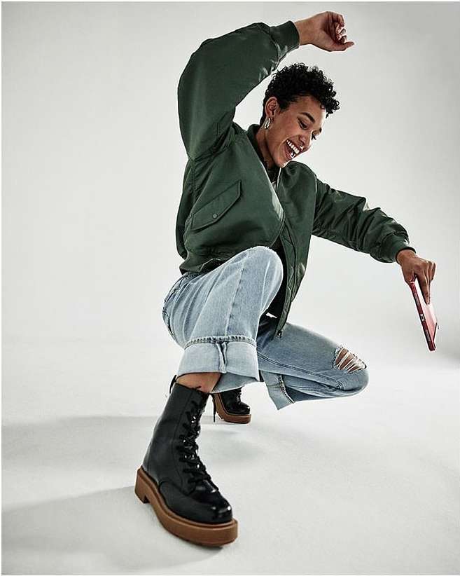 A woman in a green bomber jacket, cuffed jeans, and black boots smiles and crouches indoors, holding a tablet against a white backdrop.