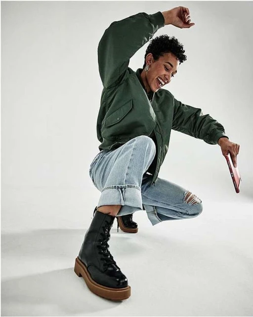 A woman in a green bomber jacket, cuffed jeans, and black boots smiles and crouches indoors, holding a tablet against a white backdrop.