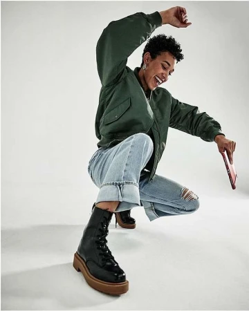 A woman in a green bomber jacket, cuffed jeans, and black boots smiles and crouches indoors, holding a tablet against a white backdrop.