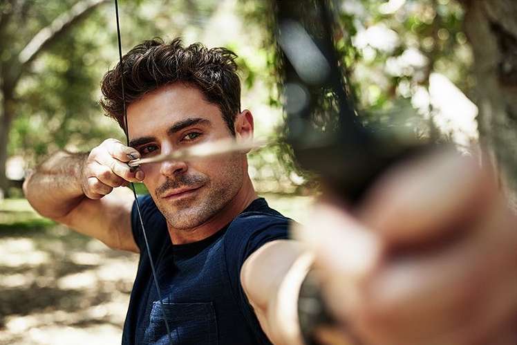 Adult male aims bow and arrow in outdoor setting. Green trees provide a natural backdrop for the active archer.