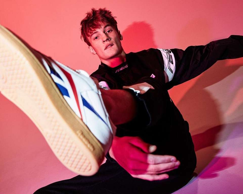 Young man in active pose, dressed in black athletic wear and sneakers with red, white, and blue detail, against a pink background.