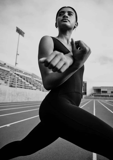 Monochrome shot of a female athlete stretching on a running track with stadium bleachers in the background on an overcast day.