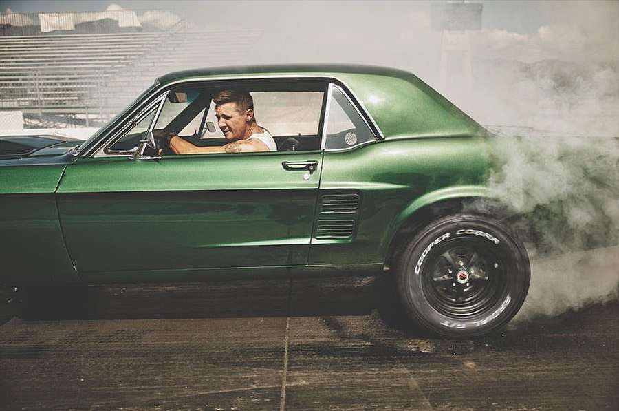 Classic green muscle car performs a burnout on the race track with a driver visible in the driver's seat and smoke coming from rear tire.