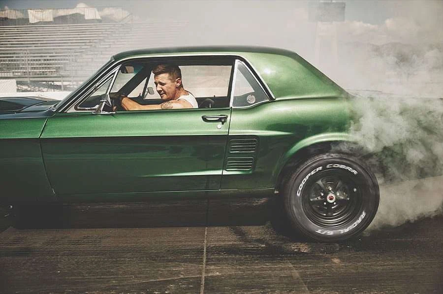 Classic green muscle car performs a burnout on the race track with a driver visible in the driver's seat and smoke coming from rear tire.