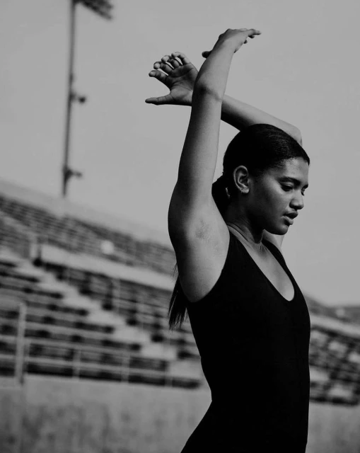 Monochrome medium shot of a woman in a black tank top stretching her arms above her head at a sports stadium.