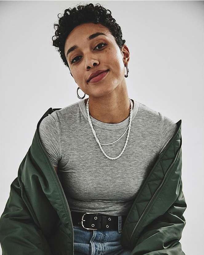 Portrait of a young adult woman with short curly hair, wearing a grey shirt, light blue jeans, silver necklaces, and an olive-green jacket.