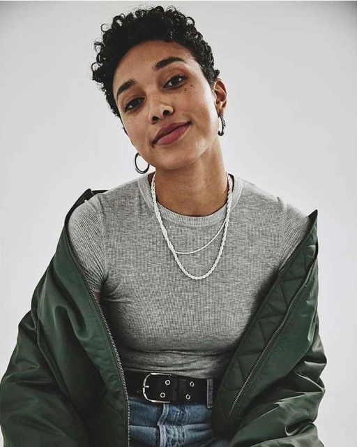 Portrait of a young adult woman with short curly hair, wearing a grey shirt, light blue jeans, silver necklaces, and an olive-green jacket.